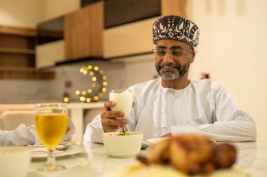 Decorations and ornaments of the month of goodness and blessings, breaking the fast by eating dates, delicious Ramadan dishes and appetizers, spending enjoyable time with family at the iftar table, an Arab Gulf Omani man wearing the dishdasha and kumma, gestures of happiness and joy, celebrating the arrival of the blessed month.