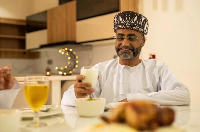 Breaking the fast with dates, delicious Ramadan dishes and appetizers, spending enjoyable time with family at the iftar table, an Arab Gulf Omani man wearing a dishdasha and kumma, gestures of happiness and joy, celebrating the arrival of the blessed month, decorations and adornments of the month of goodness and blessings.