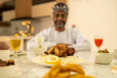 Spending quality time with family at the iftar table, an Arab Gulf Omani man wearing a dishdasha and kumma looking at the camera with gestures of happiness and joy, celebrating the arrival of the blessed month, delicious Ramadan dishes and appetizers, decorations and ornaments of the month of goodness and blessings.