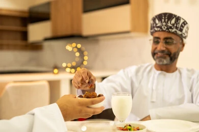 كسر الصيام بتناول التمر، مأكولات و مقبلات رمضانية شهية ، قضاء وقت ممتع مع العائلة على مائدة الافطار ، رجل عربي خليجي عماني يرتدى الدشداشة والكمة، ايماءات السعادة والسرور ، الاحتفال بقدوم الشهر المبارك ، ديكورات وزينة  شهر الخير والبركة .