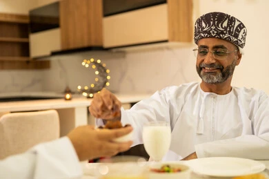 Decorations and ornaments of the month of goodness and blessings, breaking the fast by eating dates, delicious Ramadan dishes and appetizers, spending enjoyable time with family at the iftar table, an Arab Gulf Omani man wearing the dishdasha and kumma, gestures of happiness and joy, celebrating the arrival of the blessed month.