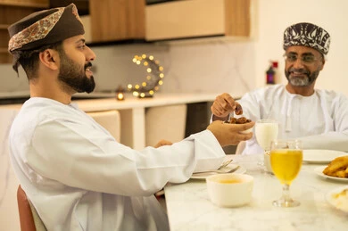 سفرة مليئة بالمقبلات والاطباق الرمضانية الشهية، لمة الاصدقاء على سفرة رمضان, صديقان عربيان خليجيان عمانيان يرتديان الدشداشة والعمامة يقومان بتناول الطعام بايماءات السرور بقدوم الشهر الفضيل, الاحتفال بالاعياد و المناسبات الاسلامية, اجتماع العائلة على المائدة