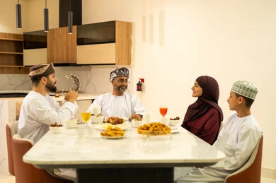 موعد كسر الصيام عند أذان المغرب في رمضان المبارك، فطور رمضاني عائلي، عائلة عربية خليجية عمانية تجلس بايماءات السرور, حسن الضيافة وإكرام الأخرين, أجواء عائلية روحانية دينية إسلامية, اجتماع الأسرة حول مائدة الإفطار المليئة بأصناف المأكولات والمقبلات الشهية