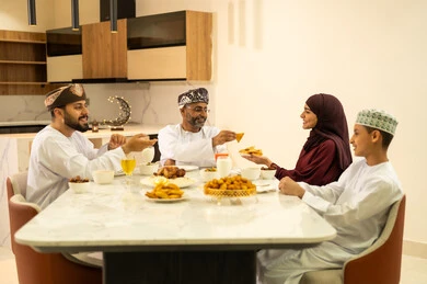 حسن الضيافة وإكرام الأخرين, موعد كسر الصيام عند أذان المغرب في رمضان المبارك، فطور رمضاني عائلي، عائلة عربية خليجية عمانية تجلس بايماءات السرور, أجواء عائلية روحانية دينية إسلامية, اجتماع الأسرة حول مائدة الإفطار المليئة بأصناف المأكولات والمقبلات الشهية