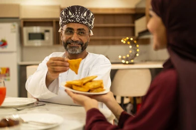 The family gathers around the iftar table filled with various delicious dishes and appetizers, the time to break the fast at the call to prayer at sunset during the month of Ramadan, decorations and accessories for the blessed month of Ramadan, a woman serving samosas to her husband, an Arab Gulf Omani family sitting with gestures of joy.