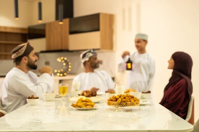 اجتماع الأسرة حول مائدة الإفطار المليئة بأصناف المأكولات والمقبلات الشهية, ولد يرتدي الكمة والدشداشة يحمل فانوس رمضان، موعد كسر الصيام عند أذان المغرب في شهر رمضان، عائلة عربية خليجية عمانية تجلس بايماءات السرور، زينة واكسسوارات شهر رمضان المبارك