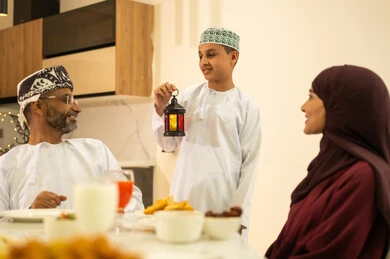 The family gathers around the iftar table filled with various delicious dishes and appetizers, a boy wearing a kumma and a dishdasha holding a Ramadan lantern, the time to break the fast at the call to prayer at sunset during the month of Ramadan, an Arab Gulf Omani family sitting with gestures of joy, decorations and accessories for the blessed month of Ramadan.