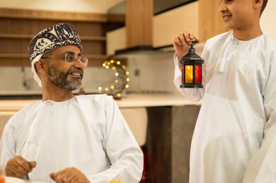 Decorations and accessories for the blessed month of Ramadan, the family gathering around the iftar table filled with various delicious dishes and appetizers, a boy wearing the kumma and dishdasha holding a Ramadan lantern, the time to break the fast at the call to prayer at sunset during Ramadan, an Arab Gulf Omani family sitting with gestures of joy.