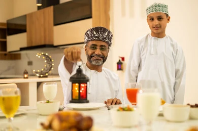 حسن الضيافة وإكرام الأخرين, موعد كسر الصيام عند أذان المغرب في رمضان المبارك، فطور رمضاني عائلي، عائلة عربية خليجية عمانية تجلس بايماءات السرور, أجواء عائلية روحانية دينية إسلامية, اجتماع الأسرة حول مائدة الإفطار المليئة بأصناف المأكولات والمقبلات الشهية