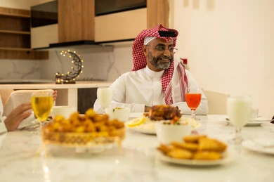 A family Ramadan breakfast, a variety of delicious dishes and appetizers, a man wearing a traditional thobe and shemagh sitting at the breakfast table with his family, the time to break the fast at the call to prayer at sunset during the blessed month of Ramadan, a Saudi Gulf Arab family sitting with gestures of joy.