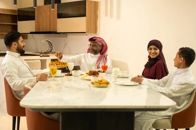 The joy of breaking the fast with family during Ramadan, a Saudi Gulf Arab family wearing traditional attire sitting at the dining table, a happy Ramadan atmosphere, a variety of foods, decorations, and accessories for the blessed month of Ramadan.