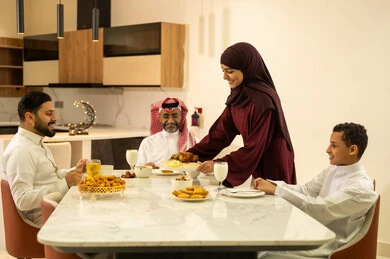 بهجة الإفطار مع العائلة في شهر رمضان  , عائلة عربية خليجية سعودية يرتدون الثوب التقليدي يجلسون على مائدة الطعام , أجواء رمضانية سعيدة, تشكيلة متنوعة من الأطعمة ، زينة واكسسوارات شهر رمضان المبارك