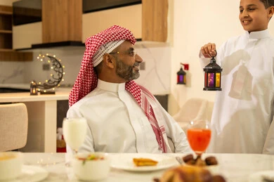 اجواء وزينة واكسسوارات رمضانية، المأكولات والمقبلات الرمضانيه المتنوعه والشهية، عائلة عربية خليجية سعودية على مائدة الافطار فى شهر رمضان الكريم في المنزل، ابن يرتدي الثوب السعودي التقليدي يحمل فانوس رمضان يقف امام والده، شهر الخير والبركة، قضاء وقت ممتع مع الأسرة