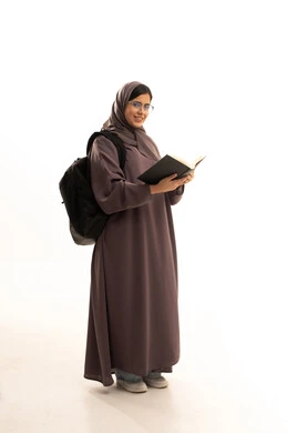 إيماءات السعادة و السرور بالمطالعة، تمضية أوقات الفراغ بالتعلم والتثقف الذاتي, بورتريه لطالبة عربية خليجية سعودية ترتدي الحجاب و العباءة و الحقيبة المدرسية تقوم بالقراءة بتمعن , مطالعة القصص التصويرية المسلية, تنمية مهارات القراءة , الاهتمام بتصفح الكتب المفيدة، الجسم بالطول الكامل، خلفية بيضاء