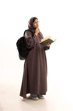 إيماءات التفكير فى شيء ما، تمضية أوقات الفراغ بالتعلم والتثقف الذاتي, بورتريه لطالبة عربية خليجية سعودية ترتدي الحجاب و العباءة و الحقيبة المدرسية تقوم بالقراءة بتمعن , مطالعة القصص التصويرية المسلية, تنمية مهارات القراءة , الاهتمام بتصفح الكتب المفيدة، الجسم بالطول الكامل، خلفية بيضاء