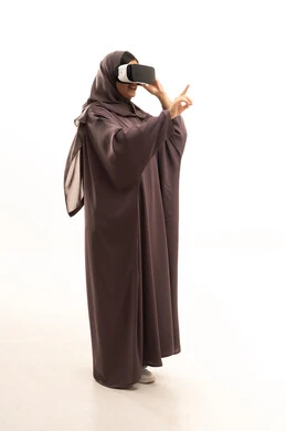 The development of technology and technology, the enjoyment and entertainment of watching through virtual reality glasses, a side portrait of a Saudi Arabian Gulf woman wearing a hijab and abaya looking through 3D glasses (VR), the integration and simulation of the imaginary world, pointing to something, a full-body image, white background.