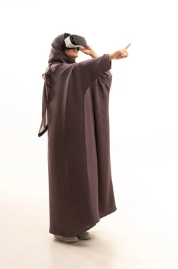 المتعة و التسلية بالمشاهدة عبر نظارة الواقع الافتراضي، تطور التقني والتكنولوجي، بورتريه من الجانب لامرأة عربية سعودية خليجية ترتدي الحجاب و العباءة تنظر من خلال نظارات ثلاثية الأبعاد (الفي ار), الاندماج ومحاكاة العالم الخيالي،الاشارة الى شي ما، صورة الجسم بالكامل، خلفية بيضاء