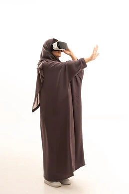 Technological and technical development, enjoyment and entertainment through watching via virtual reality glasses, a side portrait of a Saudi Arabian Gulf woman wearing a hijab and abaya looking through 3D glasses (VR), immersion and simulation of the imaginary world, pointing to something, full-body image, white background.