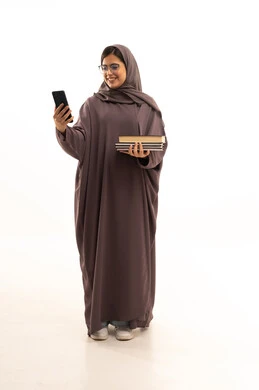 البحث والمطالعة من خلال الجوال، بورتريه لطالبة عربية خليجية سعودية ترتدي العباءة و الحجاب تستخدم الهاتف المحمول تمسك بيديها مجموعة من الكتب، تصفح التطبيقات ووسائل التواصل الاجتماعي، الجسم بالطول الكامل، خلفية بيضاء