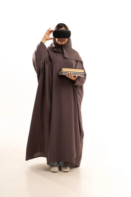 التعليم باستخدام تقنية العالم الافتراضي، دمج التقنيات الحديثة بالتعلم والتدريب، محاكاة العالم الخيالي، بورتريه  لطالبة عربية خليجية سعودية محجبة ترتدي العباءة و الحجاب تضع نظارة الفي ار ثلاثية الأبعاد تحمل مجموعة من الكتب، صورة لكامل الجسم، خلفية بيضاء