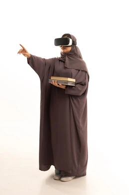 الاشارة الى شيء ما، دمج التقنيات الحديثة بالتعلم والتدريب، محاكاة العالم الخيالي، التعليم باستخدام تقنية العالم الافتراضي، بورتريه  لطالبة عربية خليجية سعودية محجبة ترتدي العباءة و الحجاب تضع نظارة الفي ار ثلاثية الأبعاد تحمل مجموعة من الكتب، صورة لكامل الجسم، خلفية بيضاء