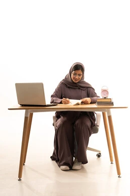 Acquiring knowledge from writing assignments, taking notes to enable and understand lessons, performing effective educational tasks, a portrait of a Gulf Arab Saudi student wearing an abaya and hijab sitting at a table in front of a laptop, white background.