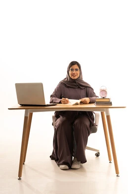 القيام بتدوين المعلومات لتمكين وفهم الدروس, اكتساب المعرفة من كتابة الواجبات، القيام بالمهام التعليمية الفعالة, بورتريه لطالبة عربية خليجية سعودية ترتدي العباءة و الحجاب تجلس على الطاولة امام الحاسب المحمول, خلفية بيضاء