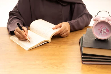 Curricula and educational materials, the concept of diligence and excellence, higher education in the Kingdom of Saudi Arabia, a close-up portrait of the hand of a Saudi Gulf Arab female student wearing an abaya, writing and reviewing daily lessons, white background.