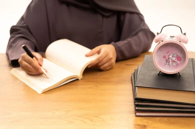 The concept of diligence and excellence, curricula and educational materials, higher education in the Kingdom of Saudi Arabia, a close-up portrait of the hand of a Saudi Gulf Arab female student wearing an abaya, writing and reviewing daily lessons, white background.