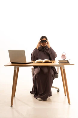 رؤية الاشياء البعيدة و تقريبها، حب الاستكشاف و المغامرة, بورتريه لطالبةعربية خليجية سعودية ترتدي الحجاب و العباءة تجلس على الطاولة امام الحاسب المحمول تحمل منظار ثنائي العينين و تنظر الى شيء ما بإيماءات السرور والاستمتاع، خلفية بيضاء