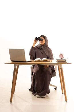 حب الاستكشاف و المغامرة, رؤية الاشياء البعيدة و تقريبها, بورتريه لطالبةعربية خليجية سعودية ترتدي الحجاب و العباءة تجلس على الطاولة امام الحاسب المحمول تحمل منظار ثنائي العينين و تنظر الى شيء ما بإيماءات السرور والاستمتاع، خلفية بيضاء