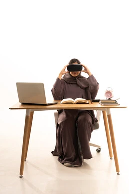 Simulation of the imaginary world, using modern technology and devices, educational platforms based on virtual reality, a portrait of a Gulf Arab Saudi female student wearing an abaya and hijab sitting at a table in front of a laptop wearing 3D VR glasses, white background.