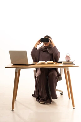 منصات تعليميه تعتمد على الواقع الافتراضي، استخدام التقنية والاجهزة الحديثة, محاكاة العالم الخيالي, بورتريه لطالبة عربية خليجية سعودية ترتدي العباءة و الحجاب تجلس على الطاولة امام الحاسب المحمول  تضع نظارة الفي ار ثلاثية الابعاد, خلفية بيضاء