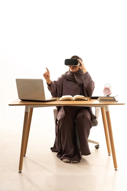 استخدام التقنية والاجهزة الحديثة، منصات تعليميه تعتمد على الواقع الافتراضي, محاكاة العالم الخيالي, بورتريه لطالبة عربية خليجية سعودية ترتدي العباءة و الحجاب تجلس على الطاولة امام الحاسب المحمول تضع نظارة الفي ار ثلاثية الابعاد, خلفية بيضاء