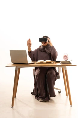 دمج الواقع الافتراضي بالتعلم والتدريب في مدارس المملكة العربية السعودية، استخدام التقنية والاجهزة الحديثة, محاكاة العالم الخيالي, بورتريه لطالبة عربية خليجية سعودية ترتدي العباءة و الحجاب تجلس على الطاولة امام الحاسب المحمول تضع نظارة الفي ار ثلاثية الابعاد, خلفية بيضاء