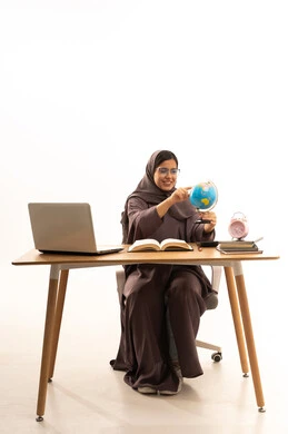 الاشارة إلى مكان معين على الخريطة , مفهوم البحث والاستكشاف حول العالم, بورتريه لطالبة عربية خليجية سعودية ترتدي العباءة تجلس على الطاولة امام الحاسب المحمول تحمل بيدها مجسم الكرة الارضية، خلفية بيضاء