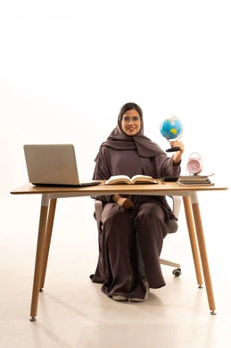 النظر إلى الكاميرا بابتسامة, مفهوم البحث والاستكشاف حول العالم, بورتريه لطالبة عربية خليجية سعودية ترتدي العباءة تجلس على الطاولة امام الحاسب المحمول تحمل بيدها مجسم الكرة الارضية، خلفية بيضاء
