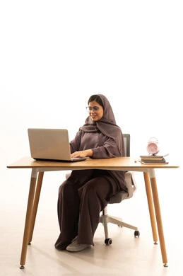 Curricula and educational materials in the Kingdom, the concept of distance education, the use of modern technological devices to follow lectures, a portrait of a university student, an Arab Gulf Saudi woman wearing a hijab, sitting at a table using a laptop for studying, white background.