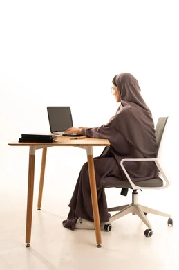 تأدية الواجبات المدرسية، استخدام الأجهزة التقنية الحديثة لمتابعة المحاضرات، المناهج والمواد التعليمية في المملكة، مفهوم التعليم عن بعد، بورتريه من الجانب لطالبة جامعية عربية خليجية سعودية محجبة تجلس على الطاولة تقوم باستخدام الحاسب المحمول فى المذاكرة، خلفية بيضاء