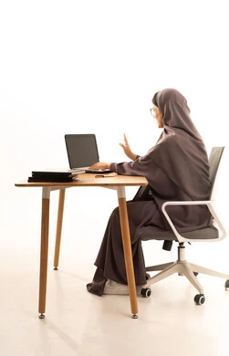 The concept of distance education, a side portrait of a Saudi Arabian Gulf hijab-wearing student in an abaya sitting at a table making a video call via a laptop, the development of science and education in Saudi Arabia, the enhancement of communication and collaboration skills through virtual education, raising a hand in gestures of peace and greeting, white background.