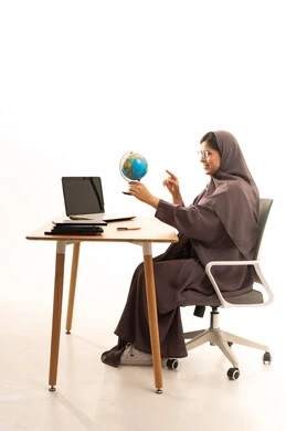 الاشارة إلى مكان معين على الخريطة، مفهوم البحث والاستكشاف حول العالم, بورتريه لإمرأة عربية خليجية سعودية ترتدي العباءة تجلس على الطاولة امام الحاسب المحمول تحمل بيدها مجسم الكرة الارضية,  خلفية بيضاء