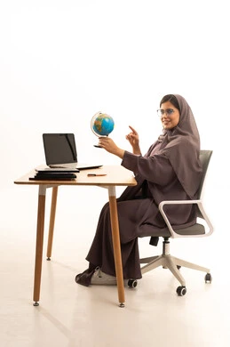 مفهوم البحث والاستكشاف حول العالم, الاشارة إلى مكان معين على الخريطة, بورتريه لإمرأة عربية خليجية سعودية ترتدي العباءة تجلس على الطاولة امام الحاسب المحمول تحمل بيدها مجسم الكرة الارضية, خلفية بيضاء
