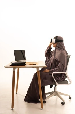 الاستمتاع و قضاء اوقات ترفيهية، التفاعل ومحاكاة العالم الافتراضي الخيالي، تقنيات حديثة الكترونية وأجهزة تكنولوجية, بورتريه لإمرأة عربية خليجية سعودية محجبة ترتدي العباءة تجلس على الطاوله امام الحاسب المحمول تضع نظارات الفي ار، خلفية بيضاء