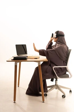 التفاعل ومحاكاة العالم الافتراضي الخيالي، تقنيات حديثة الكترونية وأجهزة تكنولوجية, بورتريه لإمرأة عربية خليجية سعودية محجبة ترتدي العباءة تجلس على الطاوله امام الحاسب المحمول تضع نظارات الفي ار، الاستمتاع و قضاء اوقات ترفيهية، خلفية بيضاء