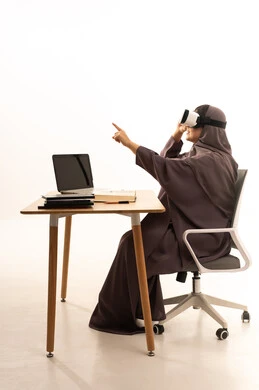 تقنيات حديثة الكترونية وأجهزة تكنولوجية، التفاعل ومحاكاة العالم الافتراضي الخيالي, بورتريه لإمرأة عربية خليجية سعودية محجبة ترتدي العباءة تجلس على الطاوله امام الحاسب المحمول تضع نظارات الفي ار،الاشارة الى شيء ما, الاستمتاع و قضاء اوقات ترفيهية، خلفية بيضاء