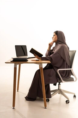 Spending free time on useful activities, gestures of thinking and contemplation, the concept of learning, excellence, and diligence, curricula and educational materials in the Kingdom of Saudi Arabia, enjoying the hobby of reading, a portrait of a Saudi Gulf Arab student wearing an abaya and hijab sitting at a table in front of a laptop reading a book, white background.