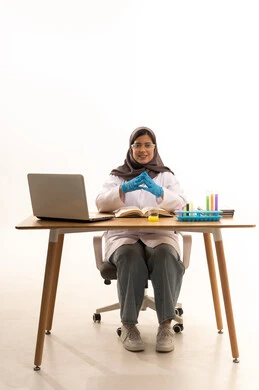 النظر الى الكاميرا بايماءات السعادة و السرور، البحث والتطوير من خلال التجارب الكيميائية المختبرية، بورتريه لطالبة عربية خليجية سعودية ترتدي المعطف الأبيض ونظارات الحماية تجلس امام الحاسوب المحمول، خلفية بيضاء