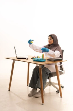 إجراء تجارب مختبرية لتطوير التجارب الكيميائية، اكتساب المعرفة والخبرة بإيماءات الدقة والتركيز، الدراسات والتجارب في بيئة عملية متطورة, بورتريه لطالبة عربية خليجية سعودية ترتدي  المعطف الأبيض ونظارات الحماية تمسك بيدها الأنبوب الزجاجي تجلس امام الحاسوب المحمول يقوم بتجربة علمية، خلفية بيضاء