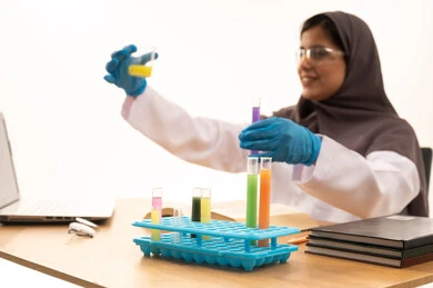 Conducting chemical experiments for scientific research and developing results, acquiring knowledge and experience with gestures of precision and focus, studies and experiments in an advanced practical environment, a close-up portrait of a Saudi Gulf Arab female student wearing a white lab coat and protective glasses holding a test tube in her hand sitting in front of a laptop conducting a scientific experiment, white background.
