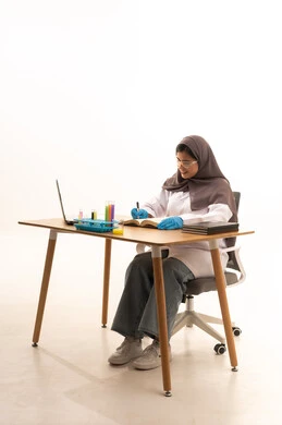 Research and development, the concept of biochemistry, conducting chemical experiments for scientific research and developing results, a portrait of a Gulf Arab Saudi female student wearing a white lab coat and safety glasses sitting in front of a laptop, taking notes and recording results, white background.